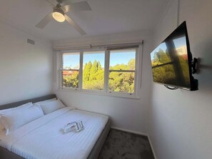 Una habitació d'hotel amb un llit, un ventilador de sostre, un televisor fixat a la paret i una finestra amb vista a arbres i edificis.