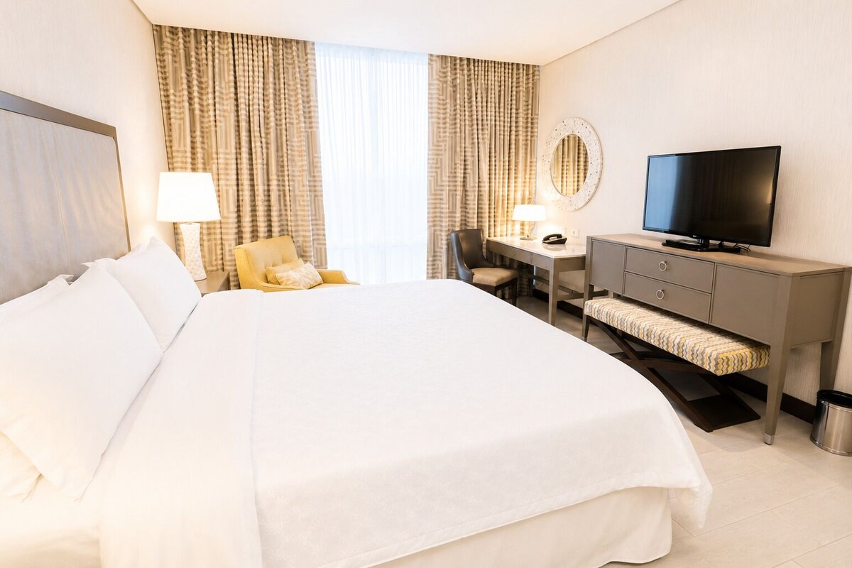 A hotel room with a large bed, a desk with a chair, a television, and a small trash bin.