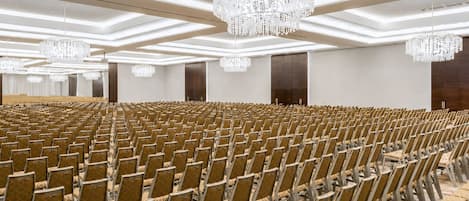 Een ruime congreszaal met rijen stoelen, een hoog plafond met kroonluchters, en een neutraal kleurenschema.