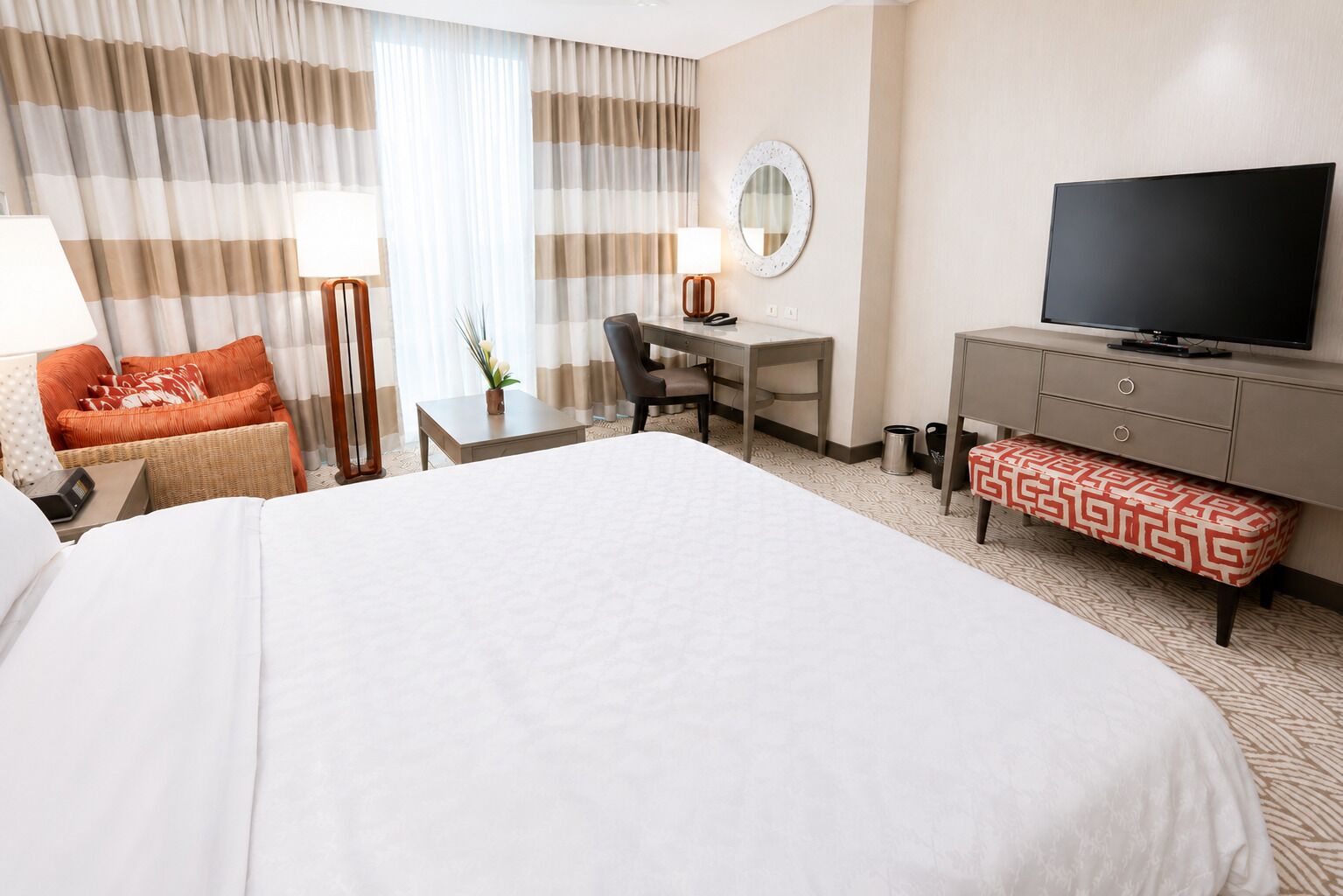 A hotel room with a large bed, a desk with a chair, a television, a small table with a lamp, and a bench.