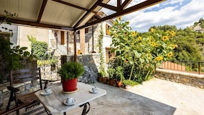 Eine Terrassengaststätte mit Tisch und Stühlen, umgeben von Topfpflanzen und Blick auf ein Steinhaus.