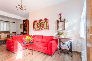 A living room with a red sofa, a small table with a vase, a sewing machine, and a colorful tapestry on the wall.