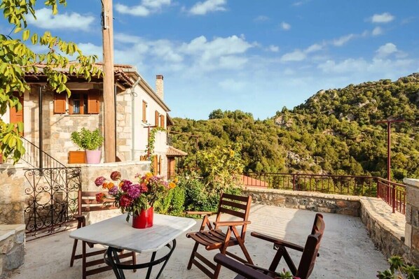 A terrace with a table and chairs, a stone building with wooden shutters, and a view of a hilly landscape.