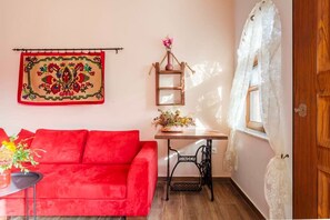 A room with a red sofa, a small table with a vase of flowers, a wooden shelf with a vase, a sewing machine, and a window with a white curtain.
