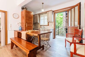 A spacious kitchen with wooden cabinets, a dining table, and a bench. There is a balcony with a view of greenery. - Stemnitsa Stone Residence - Cosy Mountain Escape (Stemnitsa)