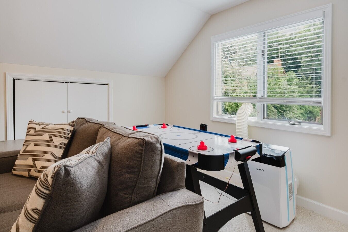 A cozy room with a foosball table, a sofa with patterned cushions, and a window with blinds.