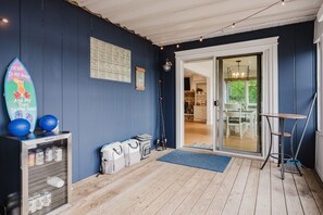 A covered outdoor area with a wooden floor, a surfboard, two basketballs, a surfboard rack, a door leading to an indoor space, and a small table with a broom.