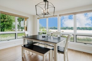 A dining area with a table and chairs, large windows with a view of greenery and a body of water, and a modern chandelier. - Wonder Lake Extended-Stay Furnished 30-Day+ Waterfront Estate (Wonder Lake)