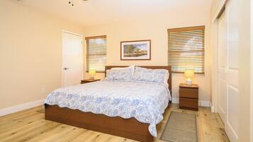 A bedroom with a wooden bed, two bedside tables with lamps, a window with blinds, and a door.