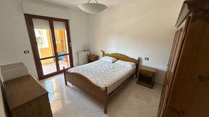 A bedroom with a wooden bed, a nightstand, a dresser, and a sliding glass door leading to an outdoor area. - House in Cagliari: Comfort, Free Parking, and Close to Brotzu Hospital (selargius)
