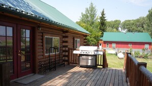 A wooden cabin with a deck, a barbecue grill, and a red building in the background. - Cozy Riverside Cabin (Riverside)