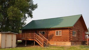 A log cabin with a green roof, a wooden deck, and a small shed. - Cozy Riverside Cabin (Riverside)