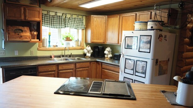 A kitchen with wooden cabinets, a built-in dishwasher, a refrigerator with magnets, and a window with a checkered valance. - Cozy Riverside Cabin (Riverside)