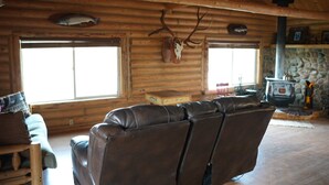 A log cabin interior with a leather sofa, a stone fireplace, and antlers on the wall. - Cozy Riverside Cabin (Riverside)