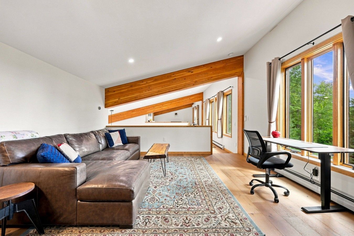 Una sala moderna con un sofá de cuero, una mesita de madera y un escritorio con una silla. Hay un ventanal con cortinas y vistas al verdor exterior.