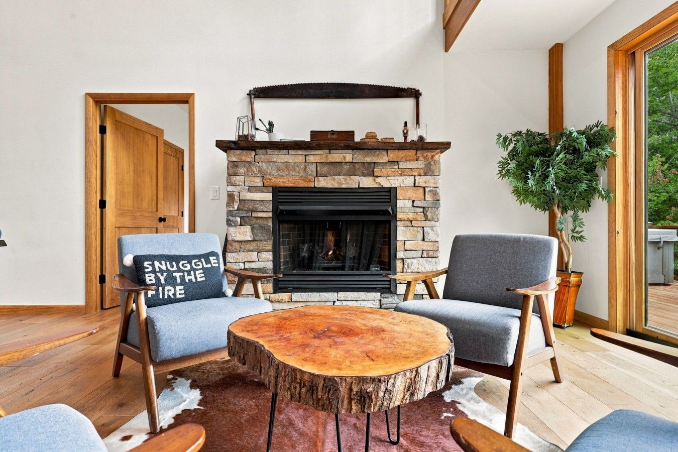 Un salon chaleureux avec une cheminée en pierre, deux fauteuils gris, une table basse ronde en bois et une grande fenêtre.