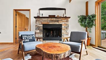 Un salon chaleureux avec une cheminée en pierre, deux fauteuils gris, une table basse ronde en bois et une grande fenêtre.