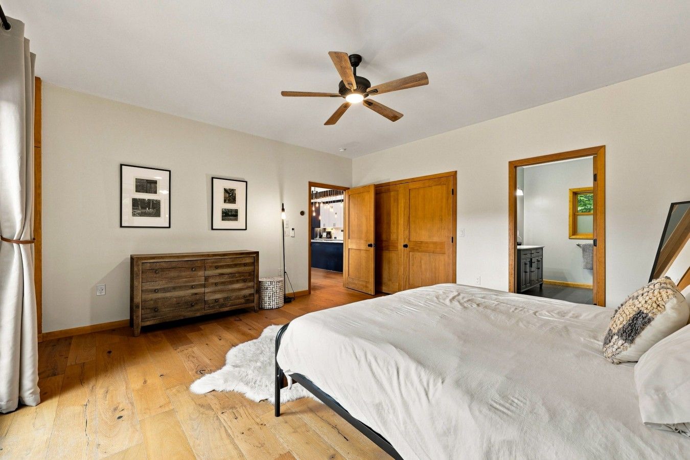 Ein Schlafzimmer mit Bett, Holzkommode, Deckenventilator, gerahmten Bildern und Blick zur Küche.