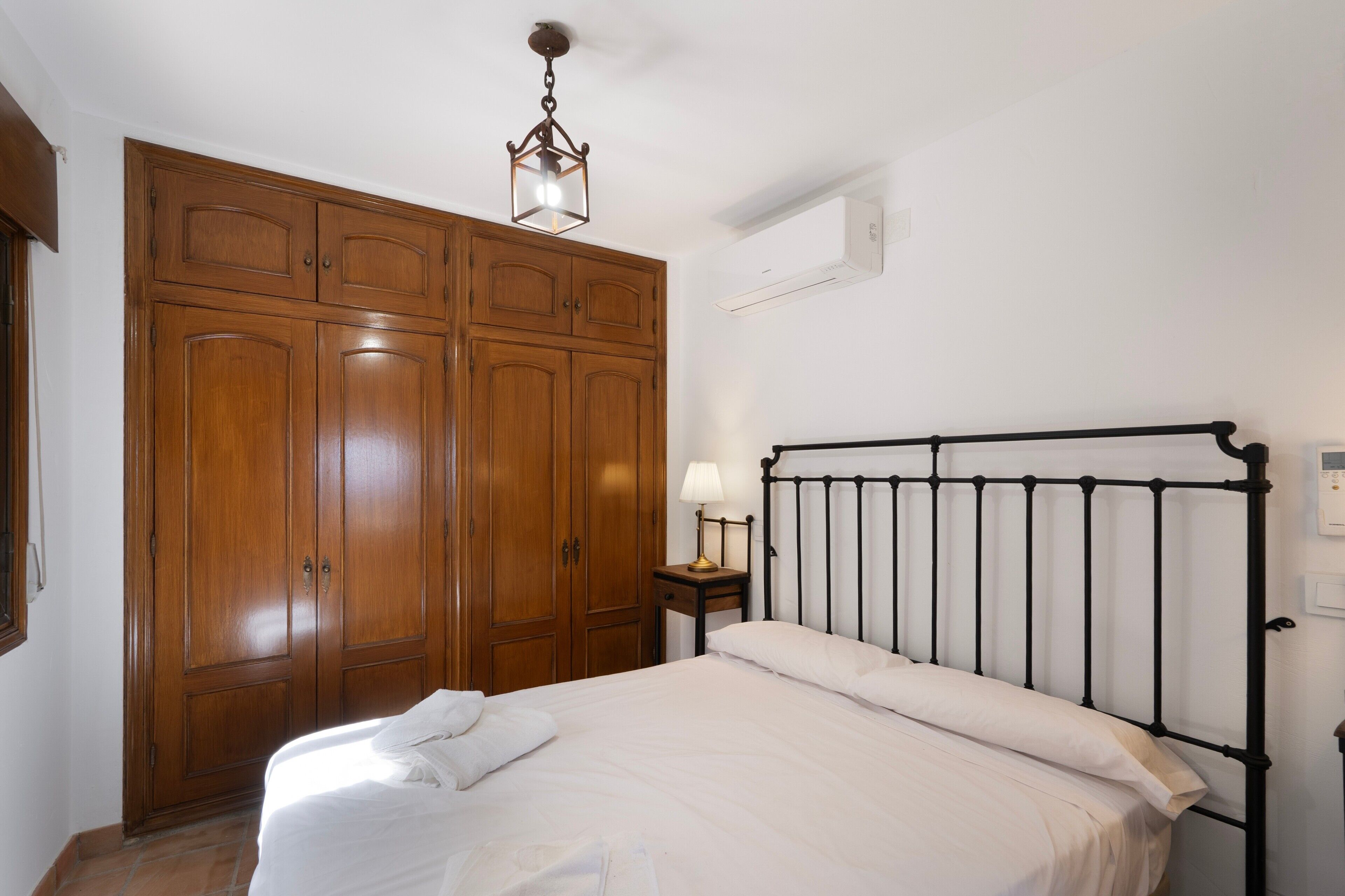 Een slaapkamer met een bed, nachtkastje, lamp, kledingkast en een plafondlamp.