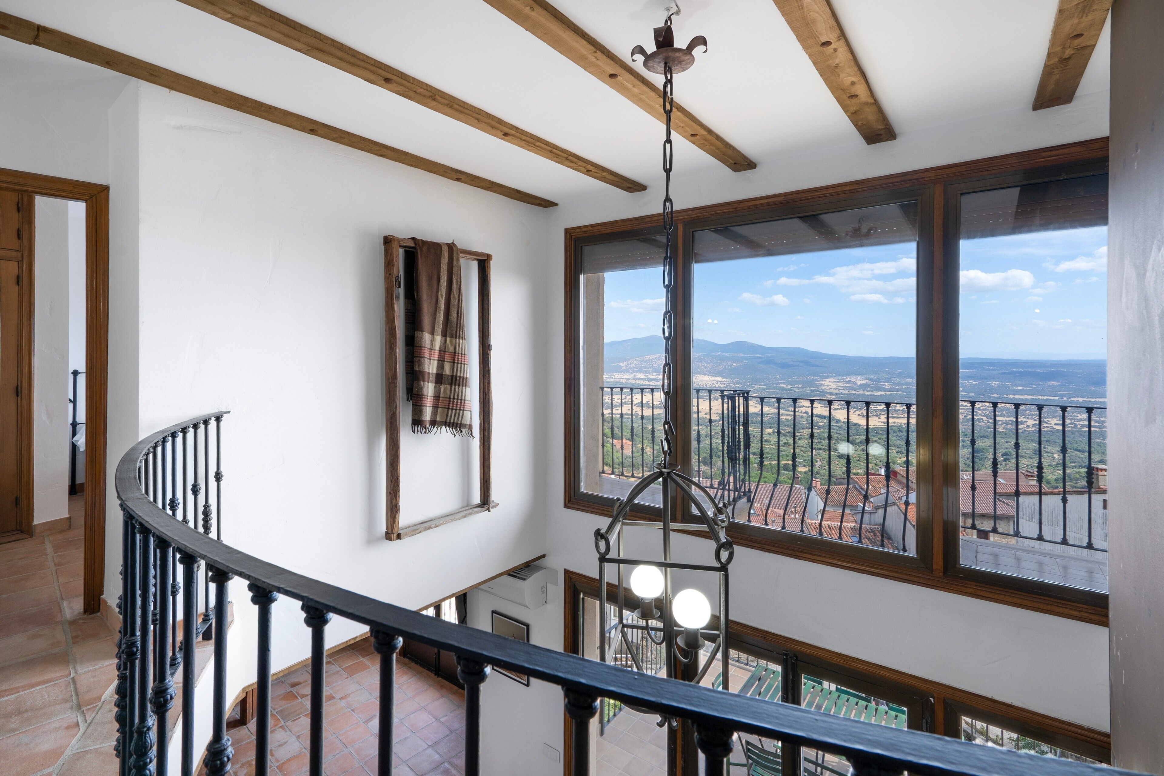 A high-ceiling room with wooden beams, a spiral staircase, and a view of a distant landscape through large windows.