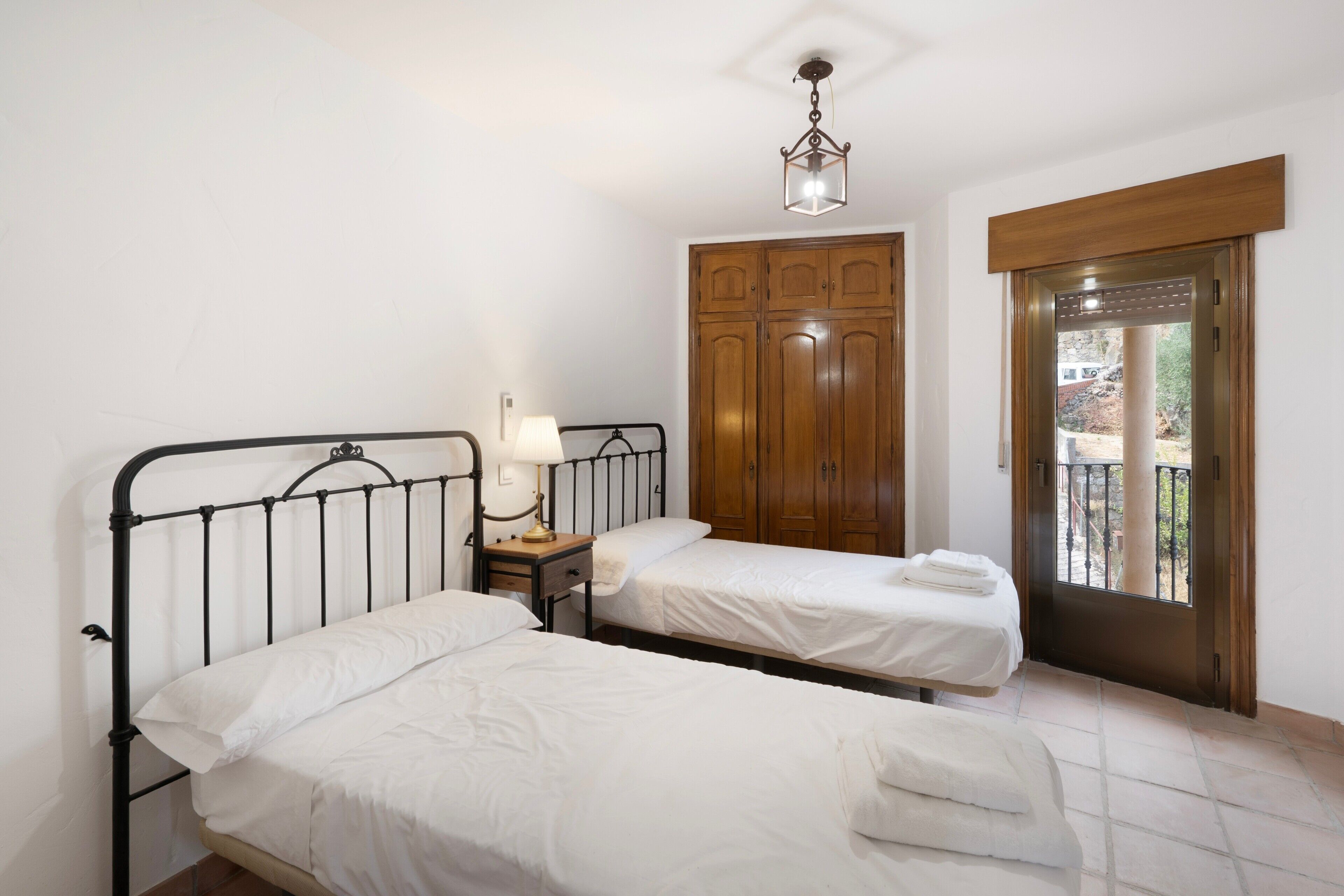 A bedroom with two single beds, a bedside table with a lamp, a wardrobe, and a door leading to an outdoor area.