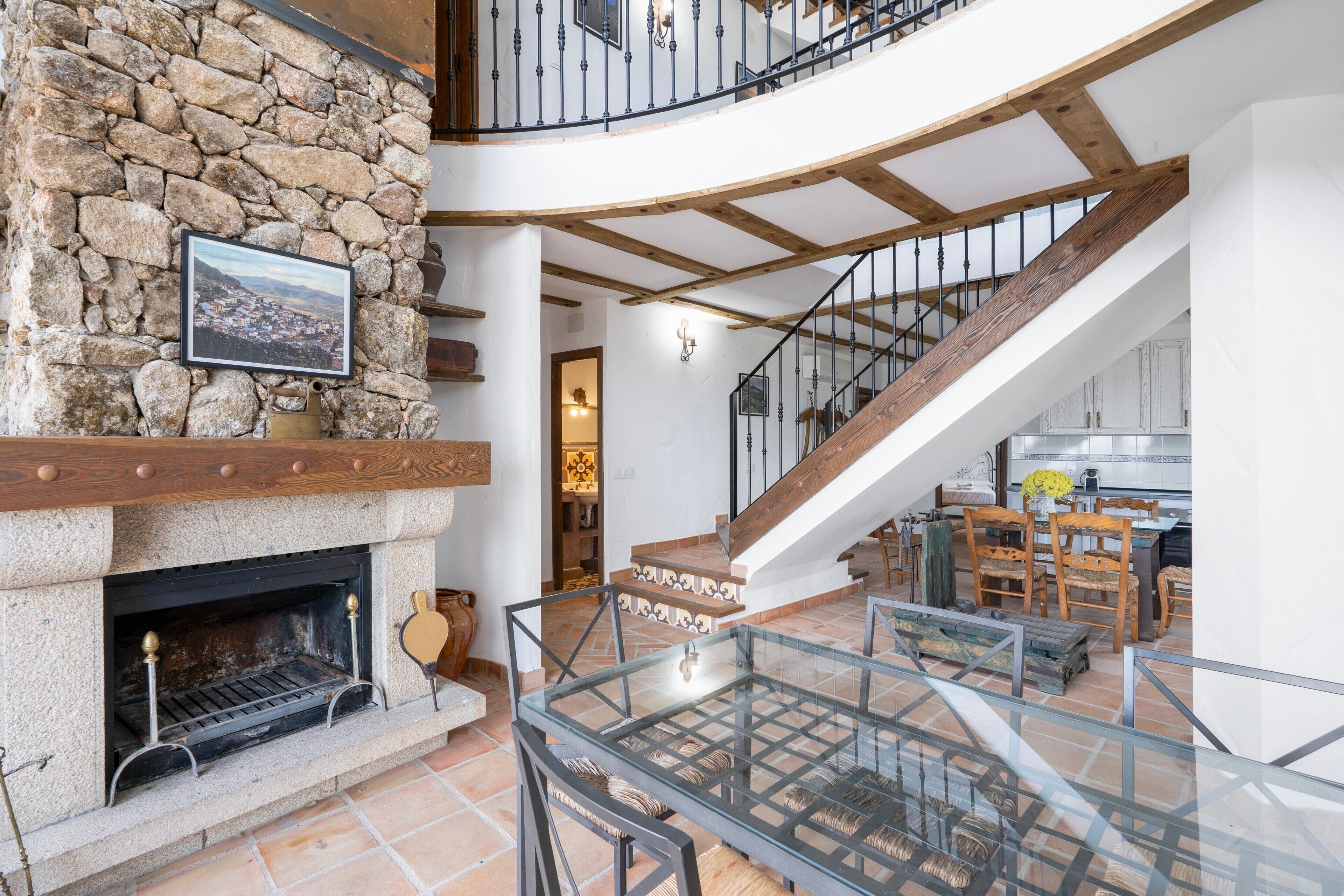 Un interior espacioso con chimenea de piedra, una escalera de madera y barandilla de cristal con vistas a un comedor.