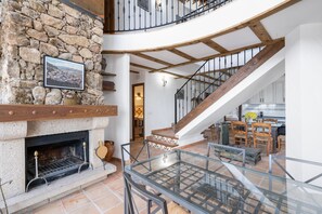 A spacious interior with a stone fireplace, a wooden staircase, and a glass railing overlooking a dining area.