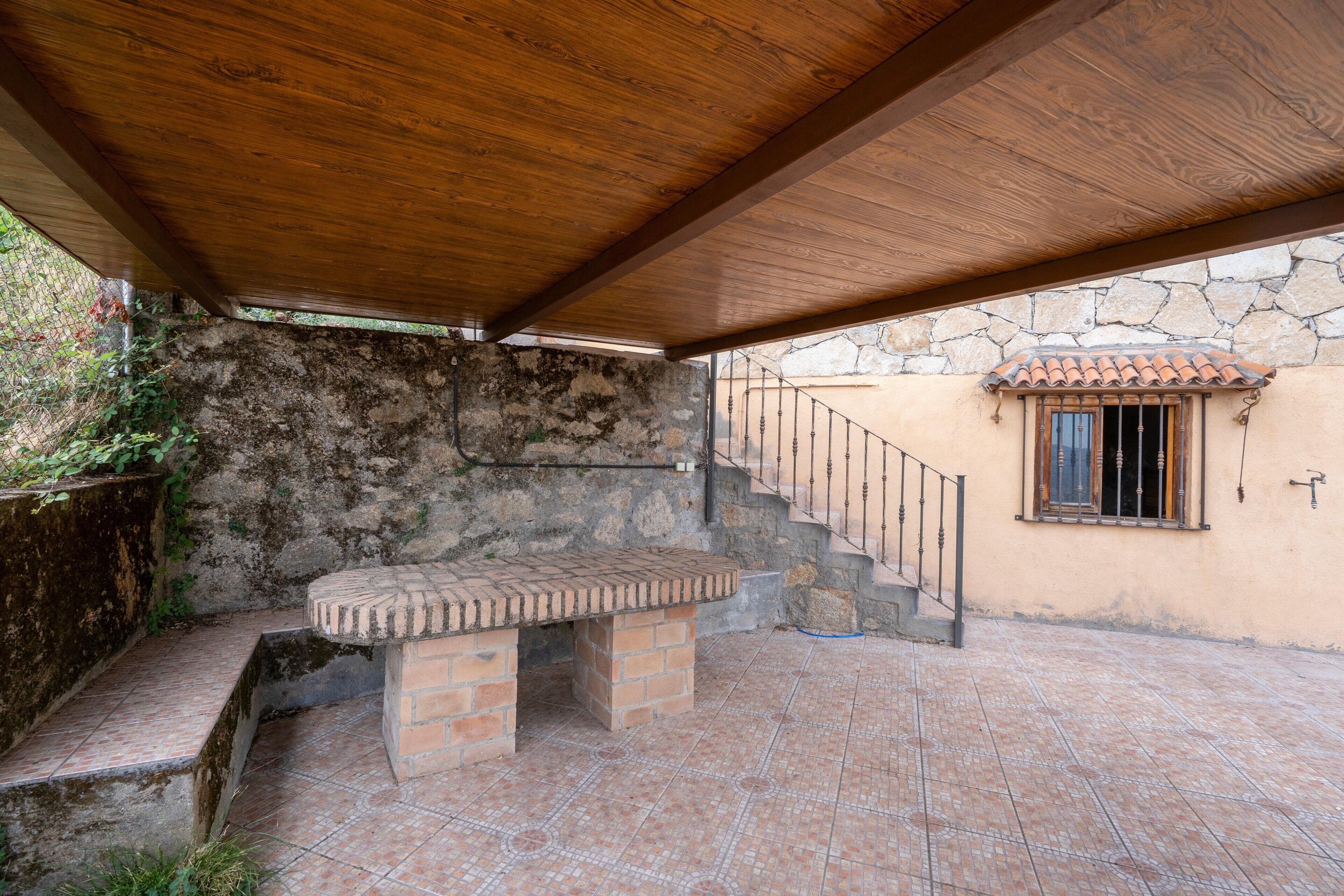 A covered patio area with a brick table, stone wall, and a staircase leading to a window.