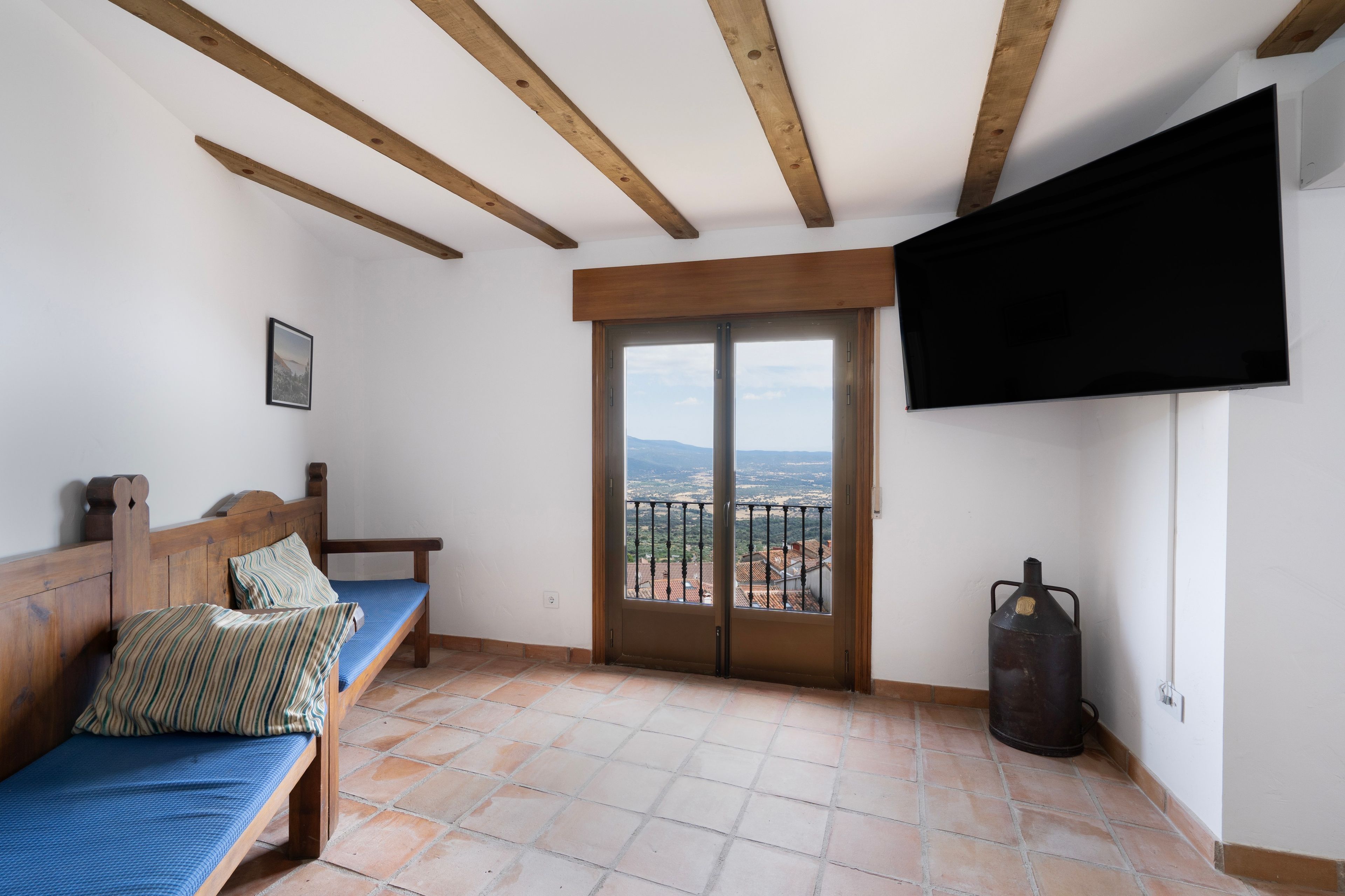 Una habitación con un banco de madera, una cama con almohada a rayas, un televisor montado en la pared y vistas a través de una puerta de cristal.