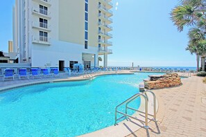 A swimming pool with blue chairs, a stone structure, and a building with balconies. - ⛱️Beachfront | 5 Pools | Majestic Beach Towers 2-407⛱️ (Panama City Beach)