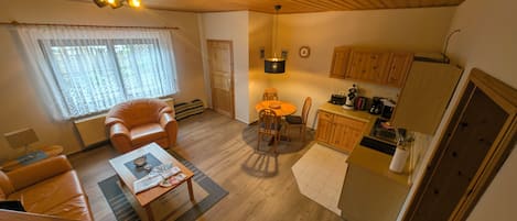 Ein gemütliches Wohnzimmer mit einer Couch, einem Couchtisch und einem Essbereich mit Stühlen. Eine Küche mit Holzschränken und Geräten ist zu sehen.