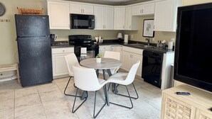 Une cuisine compacte avec des meubles blancs, un réfrigérateur noir, un four à micro-ondes et une cuisinière. On y trouve une petite table à manger avec deux chaises.