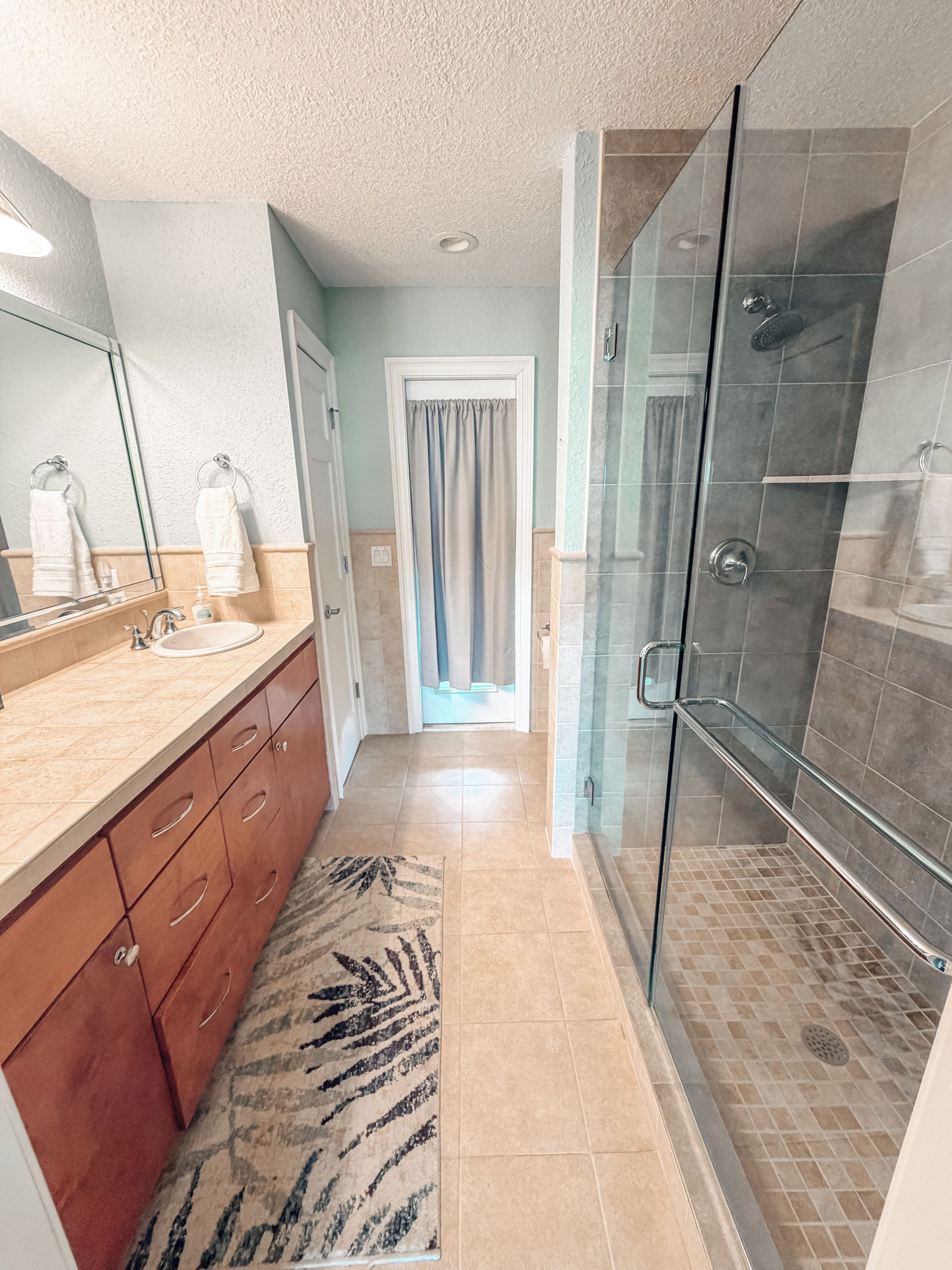 A modern bathroom with a glass shower enclosure, a sink with a mirror, and a patterned rug on the floor.