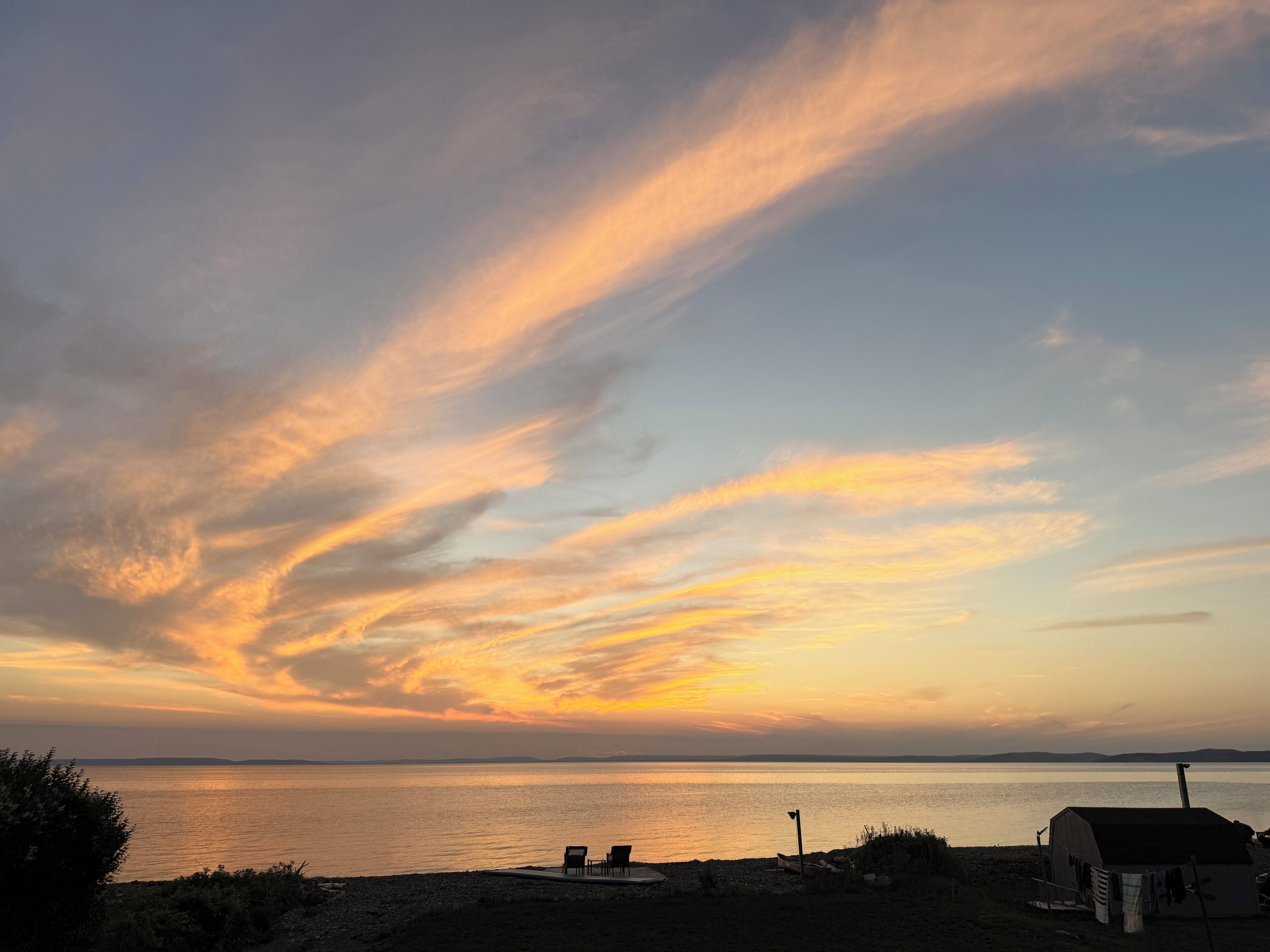Un coucher de soleil sur une étendue d’eau calme, avec une plage et un petit bâtiment.