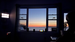 A room with a large window overlooking a sunset, a television, and a laptop on a table. - NOW BOOKING FOR 2027!! (Irish Cove)