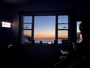 Ein Zimmer mit großem Fenster und Blick auf einen Sonnenuntergang, einem Fernseher und einem Laptop auf einem Tisch.