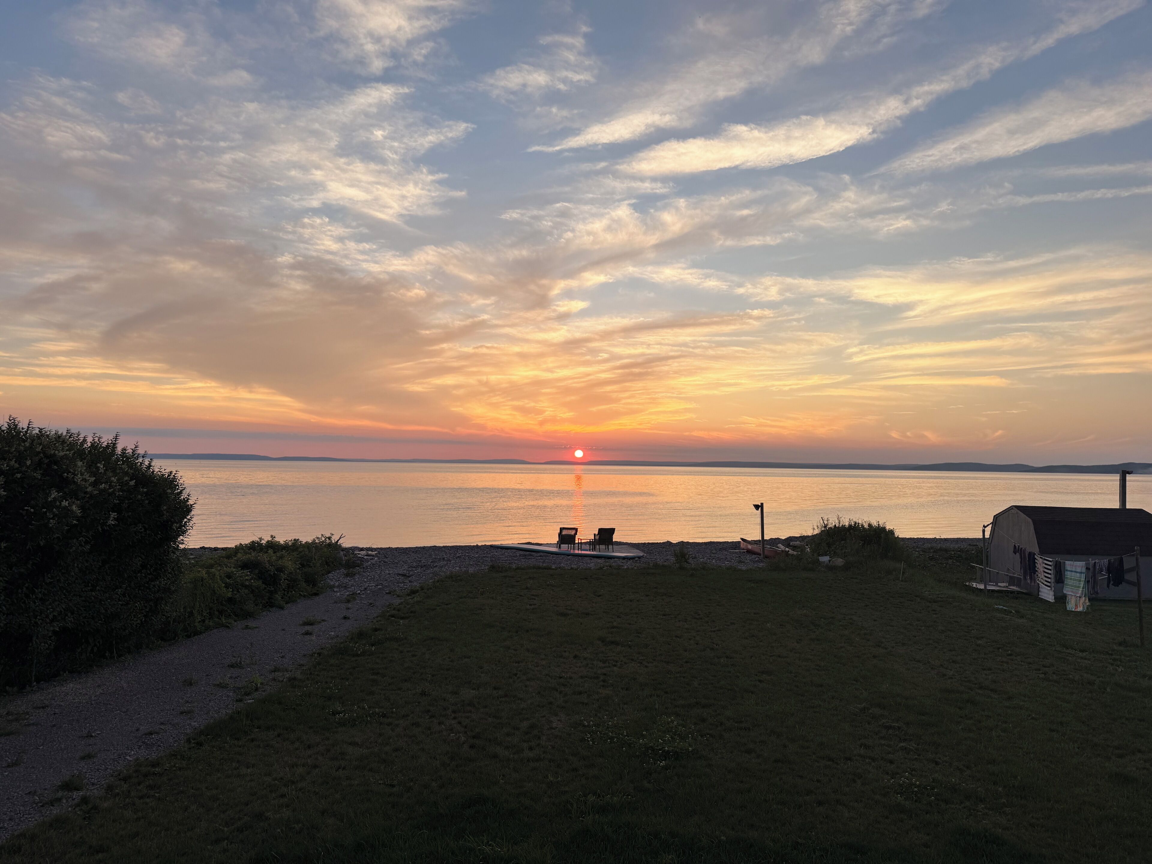 Un coucher de soleil sur une étendue d’eau calme, avec une zone herbeuse, un petit abri et quelques chaises disposées face au paysage.