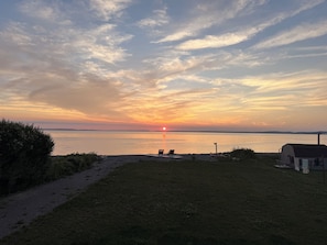 Ein Sonnenuntergang über ruhigem Wasser, mit einer Wiese, einem kleinen Schuppen und ein paar Stühlen, die die Aussicht einrahmen.