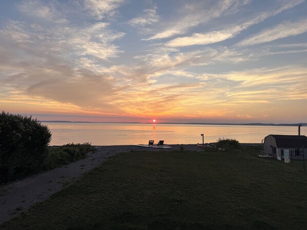 A sunset over a calm body of water with a grassy area, a small shed, and a couple of chairs facing the view.
