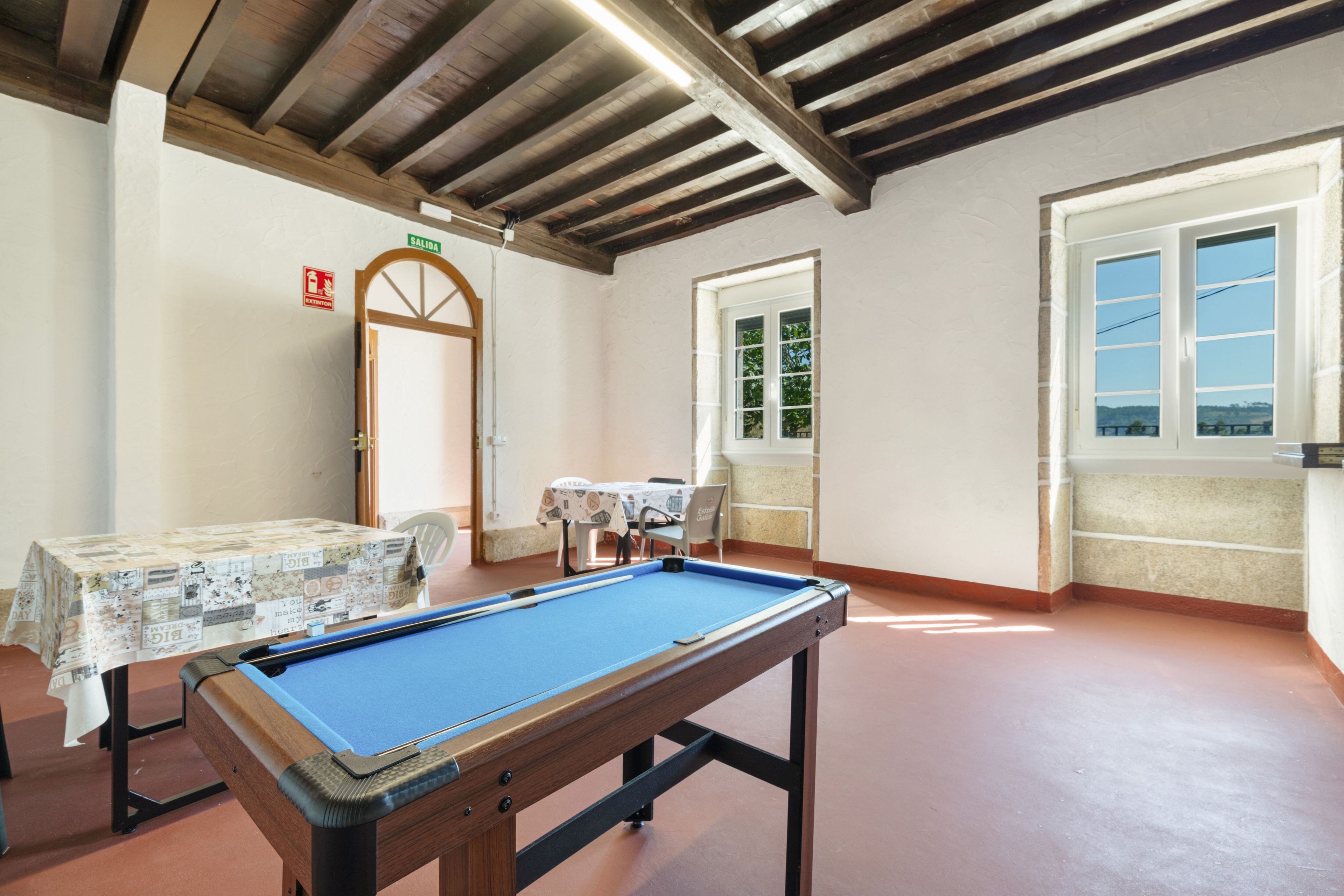 Una mesa de billar en una habitación con techo de madera, una puerta con la leyenda 'SALIDA' y una ventana con vistas.