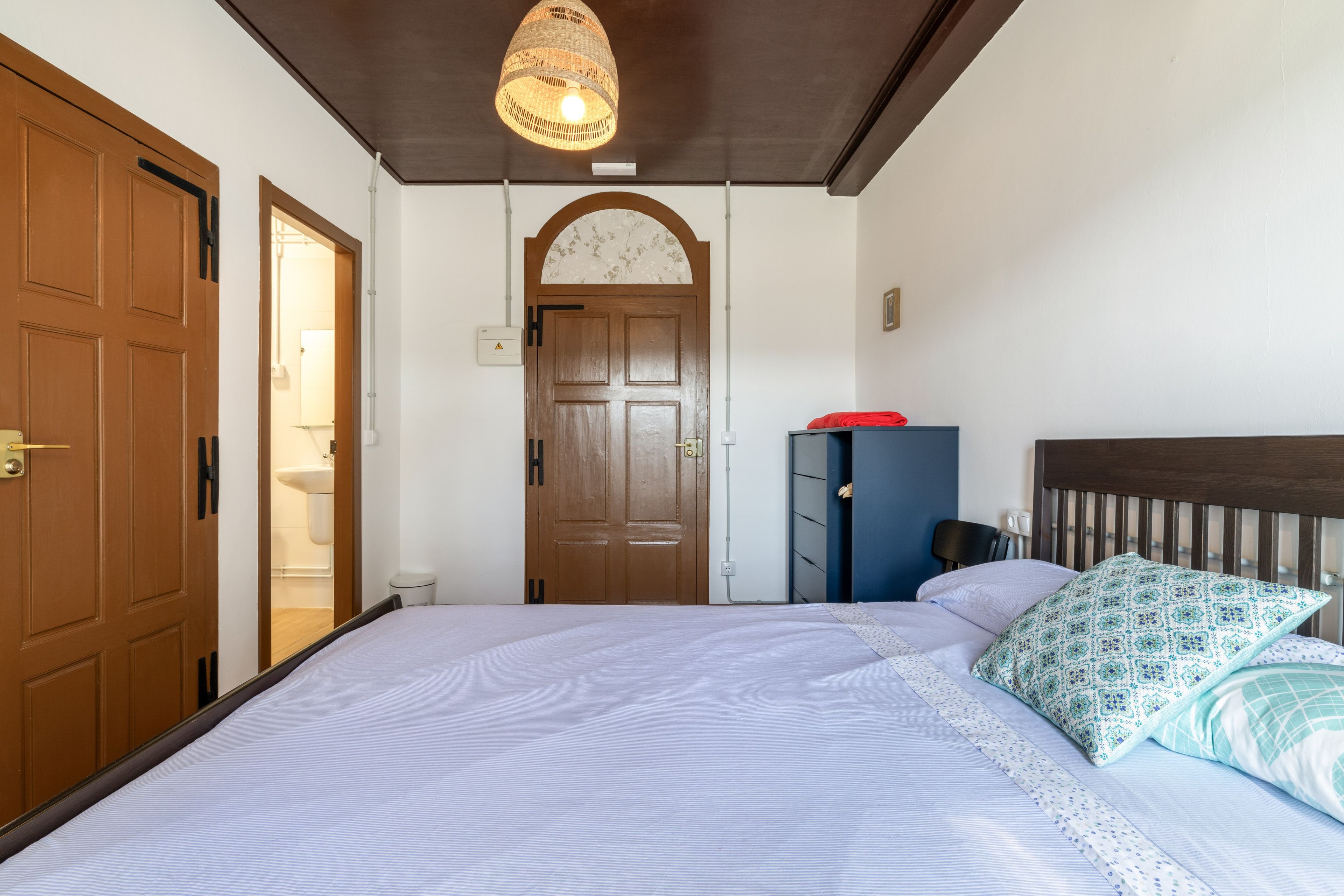 A bedroom with a large bed, a wooden wardrobe, a door leading to a bathroom, and a hanging light fixture.