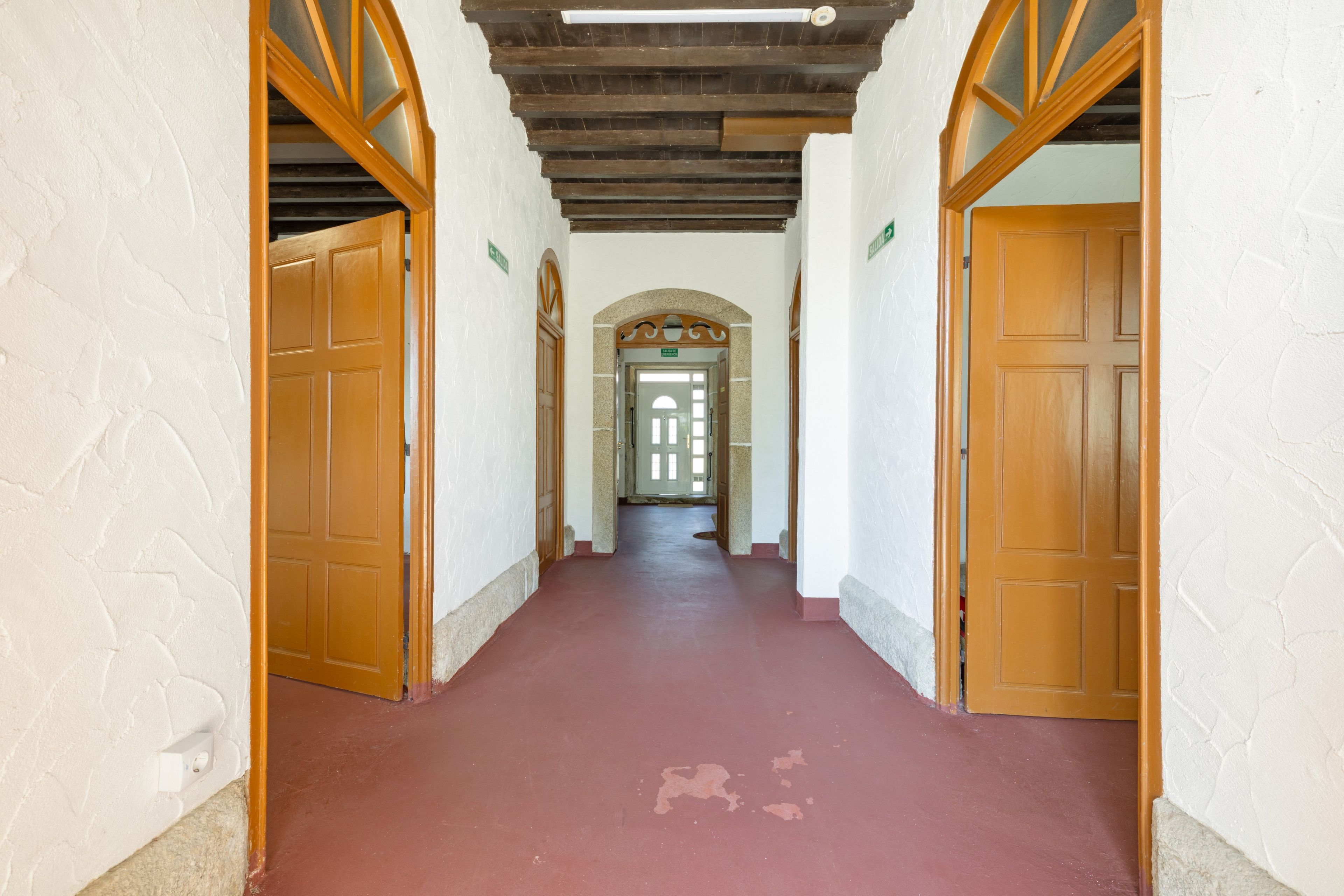 Un pasillo con puertas de madera y suelo rojo, que conduce a un acceso con panel de vidrio.
