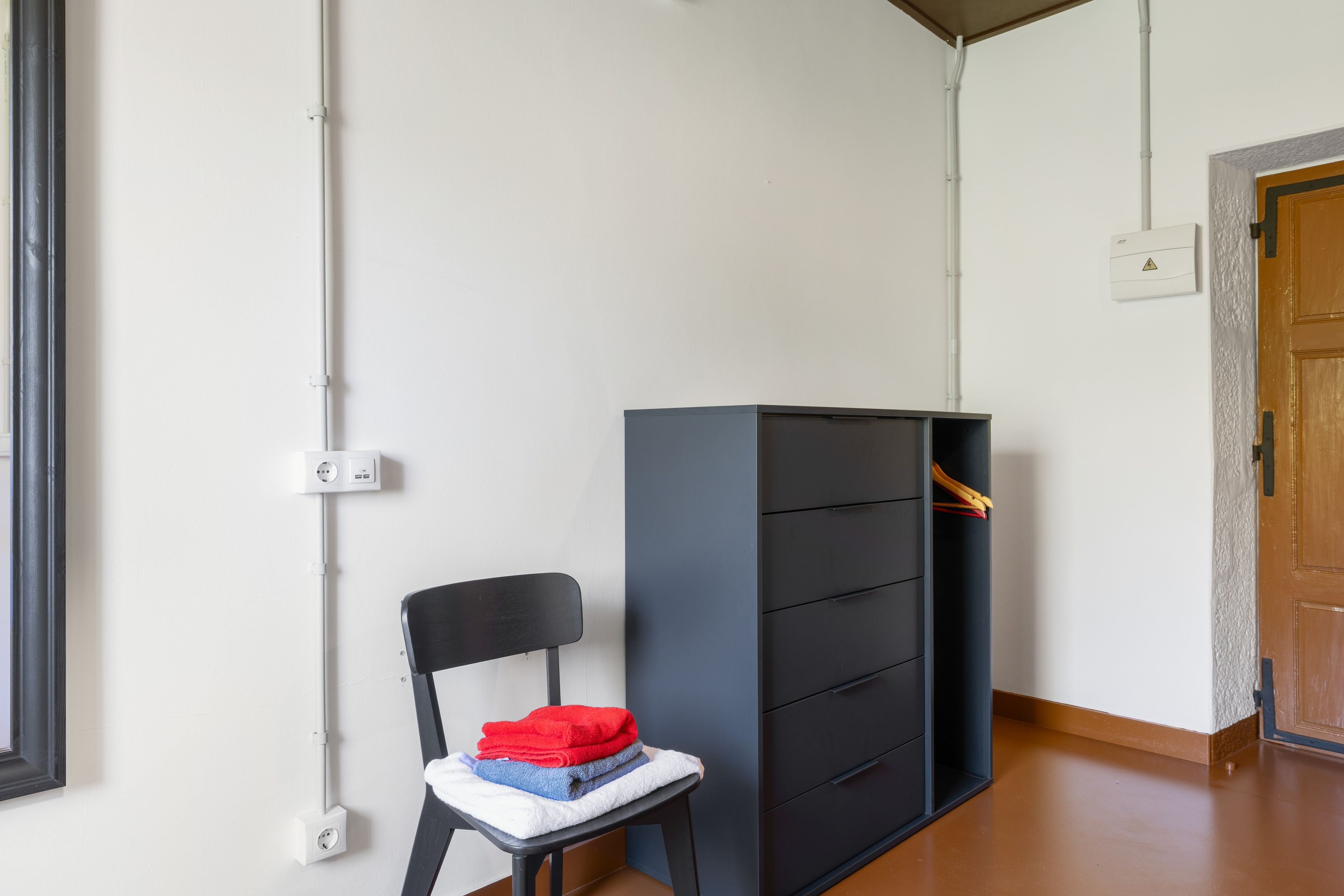 Ein Zimmer mit einem schwarzen Stuhl, einem grauen Schrank mit Schubladen, einer Holztür und einer weißen Wand mit Steckdose.