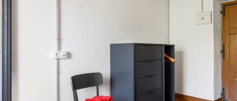 Ein Zimmer mit einem schwarzen Stuhl, einem grauen Schrank mit Schubladen, einer Holztür und einer weißen Wand mit Steckdose.