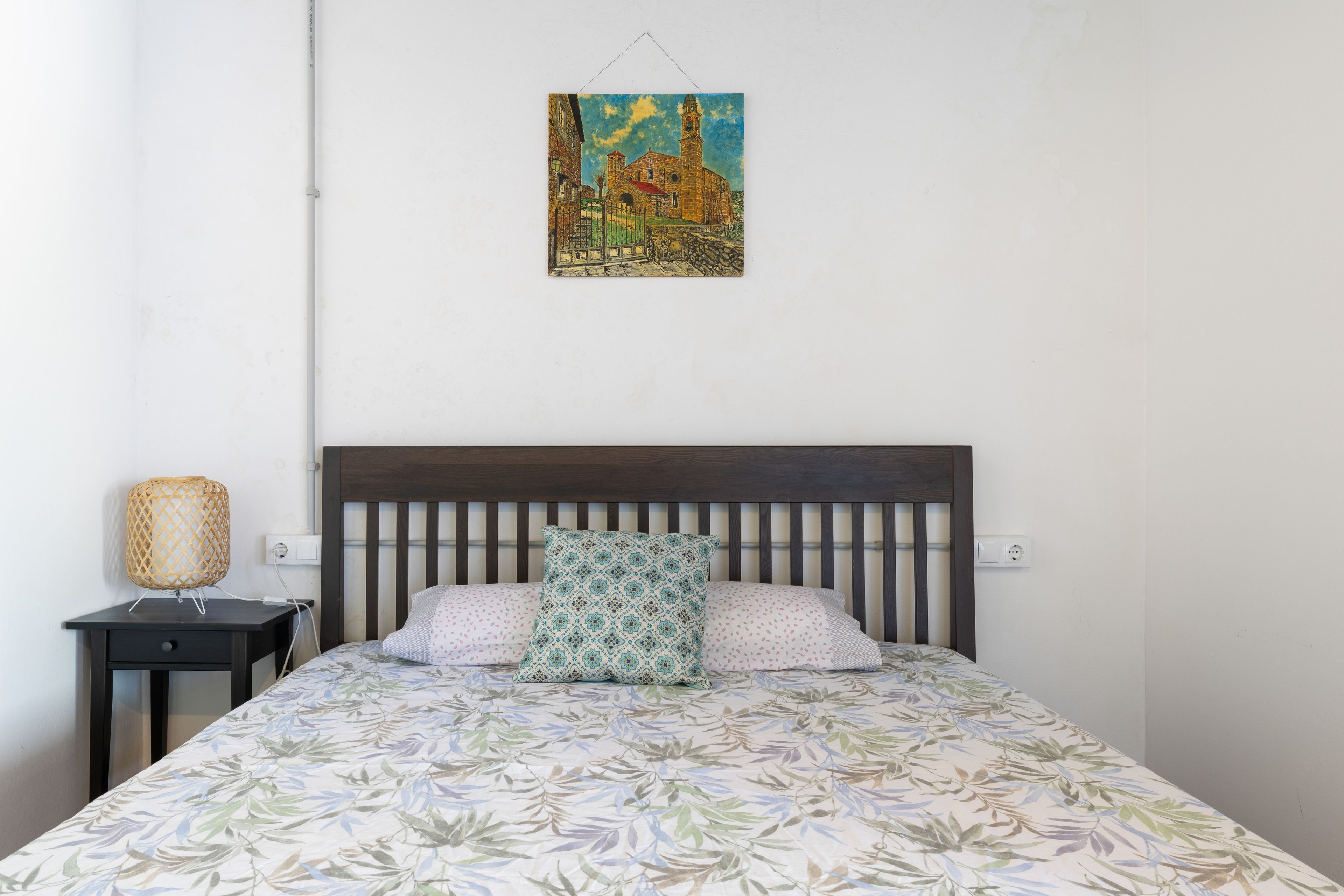 Un dormitorio con cama, mesita de noche, lámpara y un cuadro en la pared.