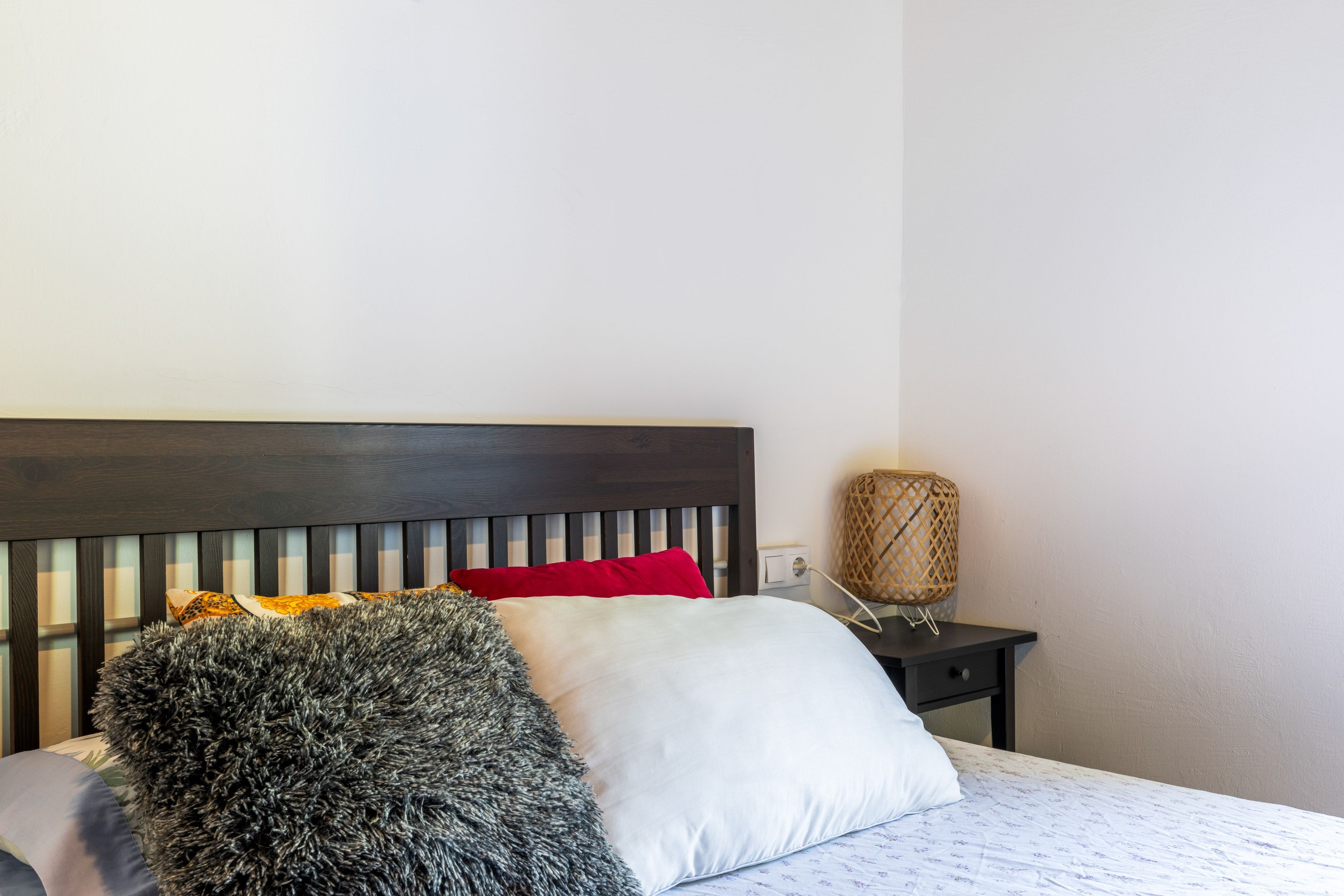 Una cama con cabecera oscura, almohadas blancas y rojas, una manta gris mullida y una mesita de noche con una lámpara de fibra.