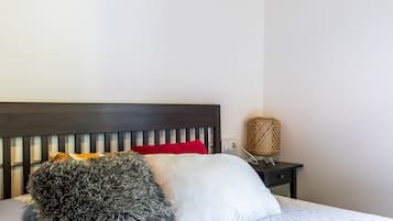 Una cama con cabecera oscura, almohadas blancas y rojas, una manta gris mullida y una mesita de noche con una lámpara de fibra.