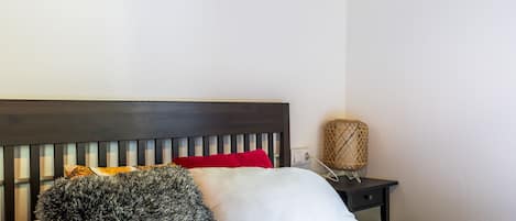 Ein Bett mit dunkelbrauner Kopfteile, weißen und roten Kissen, einer flauschigen grauen Decke und einem Nachttisch mit einer gewobenen Lampe.