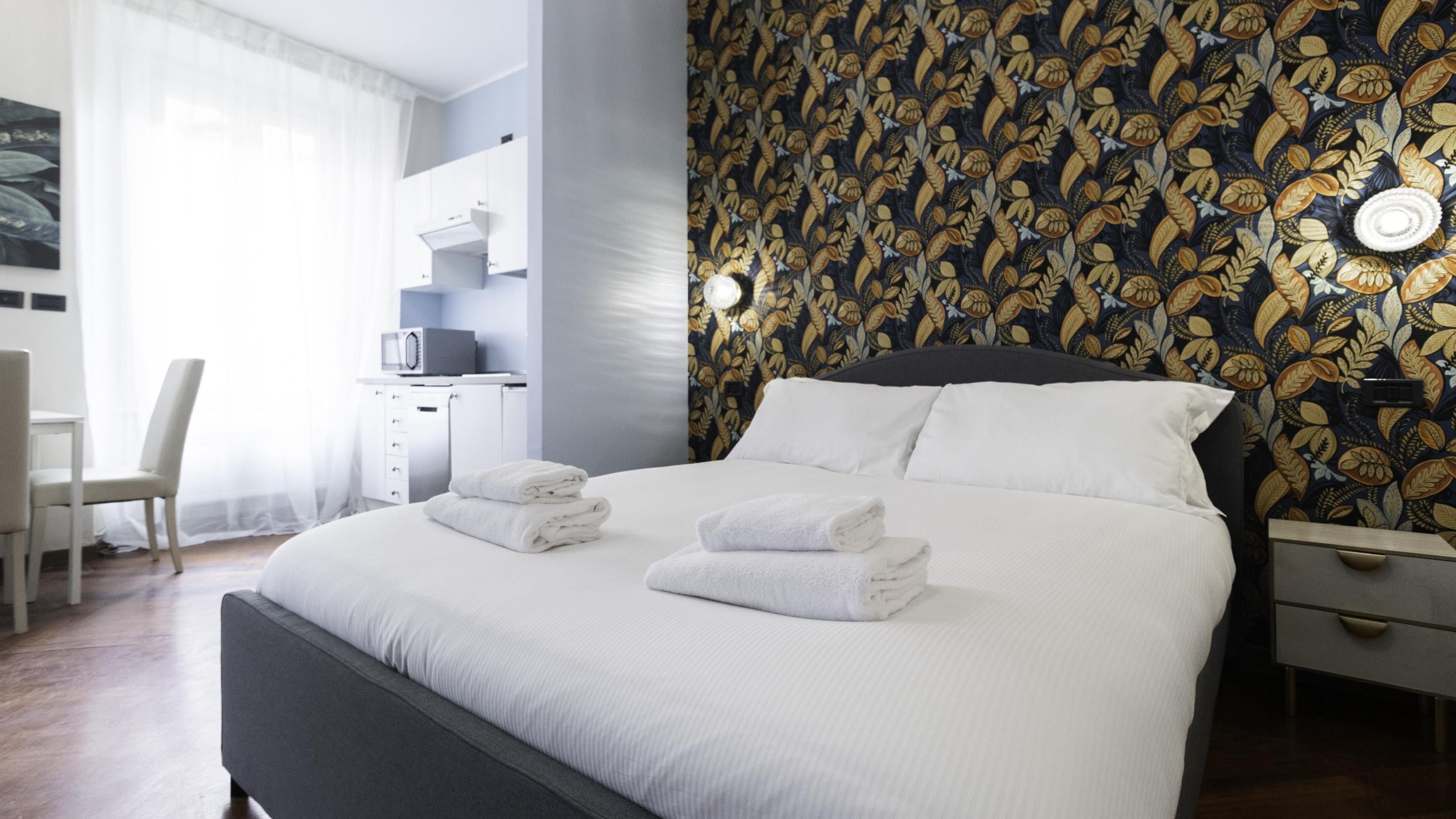 A modern hotel room with a large bed, a desk with a computer, and a patterned wallpaper.