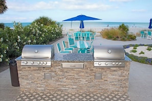 Un área de barbacoa al aire libre con dos parrillas, rodeada de sillas turquesa y una sombrilla azul, con vistas a la playa.